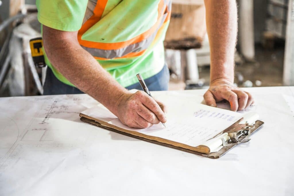 A construction worker in a high-visibility vest taking notes on a clipboard resting on top of architectural blueprints.